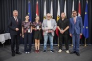 Landeshauptmann Mag. Thomas Stelzer und Wirtschafts- und Sport-Landesrat Markus Achleitner gratulieren dem ASKÖ Linz AG Froschberg. Sechs Personen stehen vor vielen Flaggen und halten Urkunden in der Hand.