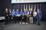Landeshauptmann Mag. Thomas Stelzer und Wirtschafts- und Sport-Landesrat Markus Achleitner gratulieren der SPG Felbermayr Wels. Viele Personen aus einer Sportmannschaft stehen vor einer Wand und halten Auszeichnungen in der Hand.