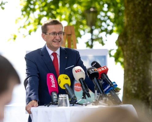 Ein Herr steht vor einem Tisch mit vielen Mikrofonen und spricht.