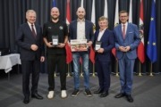 Landeshauptmann Mag. Thomas Stelzer und Wirtschafts- und Sport-Landesrat Markus Achleitner gratulieren den Raiffeisen Flyers Wels. Fünf Männer stehen vor vielen Flaggen und lächeln in die Kamera.
