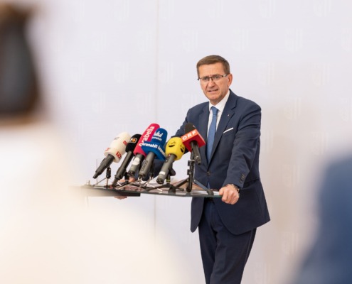 Ein Herr steht an einem Pult mir vielen Mikrofonen und spricht ins Publikum.