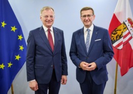 Zwei Herren stehen im Anzug vor einer Wand mit Flaggen.