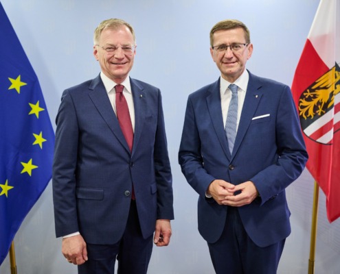 Zwei Herren stehen im Anzug vor einer Wand mit Flaggen.