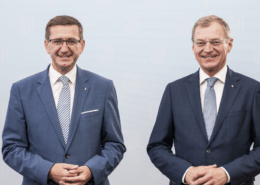 Zwei Herren stehen elegant gekleidet vor einer Wand und schauen in die Kamera.