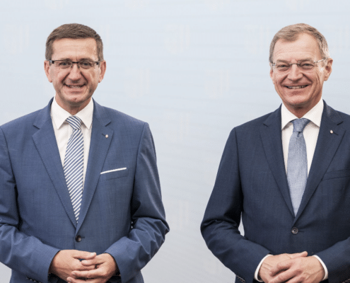 Zwei Herren stehen elegant gekleidet vor einer Wand und schauen in die Kamera.