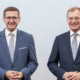 Zwei Herren stehen elegant gekleidet vor einer Wand und schauen in die Kamera.