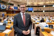 Wirtschafts- und Europa-Landesrat Markus Achleitner im Ausschuss der Regionen in Brüssel. Landesrat Markus Achleitner steht im Plenarsaal des Europäischen Parlaments. Im Hintergrund sind die Sitzplätze der Abgeordneten, Laptops und die Flaggen der EU-Mitgliedstaaten zu sehen.