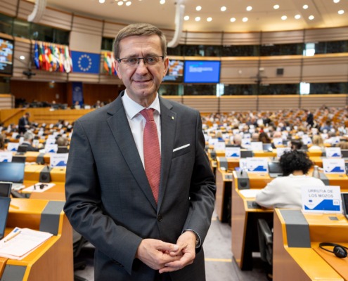 Landesrat Markus Achleitner steht im Plenarsaal des Europäischen Parlaments. Im Hintergrund sind die Sitzplätze der Abgeordneten, Laptops und die Flaggen der EU-Mitgliedstaaten zu sehen.