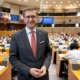 Landesrat Markus Achleitner steht im Plenarsaal des Europäischen Parlaments. Im Hintergrund sind die Sitzplätze der Abgeordneten, Laptops und die Flaggen der EU-Mitgliedstaaten zu sehen.
