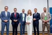 Eine Gruppe von sechs Personen steht gemeinsam für ein Foto vor einer hellen Wand. Die Personen tragen Businesskleidung und lächeln in die Kamera.