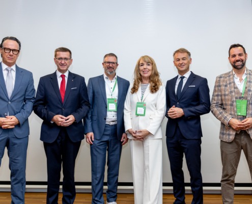 Eine Gruppe von sechs Personen steht gemeinsam für ein Foto vor einer hellen Wand. Die Personen tragen Businesskleidung und lächeln in die Kamera.