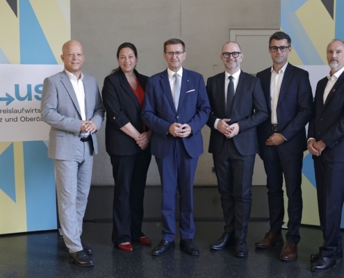 Sechs Personen, fünf Männer und eine Frau, stehen nebeneinander für ein Gruppenfoto vor einer Wand mit blau-gelben Grafikelementen und einem Banner zur Initiative „re-use“. Sie tragen Businesskleidung und blicken in die Kamera.