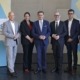 Sechs Personen, fünf Männer und eine Frau, stehen nebeneinander für ein Gruppenfoto vor einer Wand mit blau-gelben Grafikelementen und einem Banner zur Initiative „re-use“. Sie tragen Businesskleidung und blicken in die Kamera.