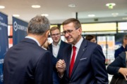 Landesrat Markus Achleitner unterhält sich mit einem Mann im Anzug bei einer Veranstaltung mit Messestand im Hintergrund.