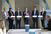 Sechs Personen stehen bei einer Pressekonferenz hinter einem langen Stehtisch mit Namensschildern und Mikrofonen. Ein Mann in blauem Anzug spricht in die Runde, während die anderen Teilnehmenden zuhören. Im Vordergrund sind unscharf Teile des Publikums und Tische zu sehen. Im Hintergrund stehen Banner zur Initiative „re-use“.