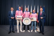 Fünf Personen stehen vor einem Vorhang mit Flaggen und halten einen Preis in der Hand.